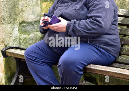 Ein FAT- oder übergewichtige Person sitzen auf einer Bank von SMS-Nachrichten oder Nachrichten mit einem Smartphone oder tragbaren Gerät. große Mann oder Frau sitzt Wiedergabe mit Telefon. Stockfoto