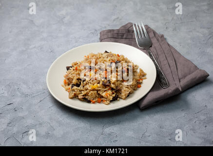 Risotto mit Pilzen und Gemüse auf einer Keramikplatte, Gabel, Serviette auf einem grauen konkreten Hintergrund Stockfoto