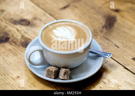 Ein frisch gebrauten Latte Kaffee in eine Tasse und Untertasse mit zwei Klumpen von braunem Zucker. Auf einem Tisch die Milch Kunstwerk in Form einer Tulpe gebildet wird gesetzt Stockfoto