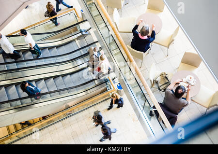 Blick von oben auf die Menschen in einem Einkaufszentrum unter den Rolltreppen, schlendern entlang der Gehwege und einen Snack auf der Terrasse eines Cafés. Stockfoto