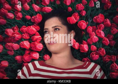 Nahaufnahme Portrait von junge schöne Mädchen Frau mit rot-braune Haare liegen auf Gras mit Rote Tulpe Blumen rund um den Kopf. Blick von oben Top overhea Stockfoto