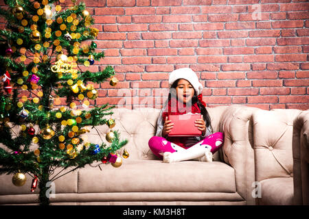 Kleine indische Mädchen, dass Weihnachten Geschenke in roten Tüchern oder Santa Clause hat und Kleidung, mit weissem oder blauen Hintergrund oder in Zeichnung isoliert stehende Stockfoto