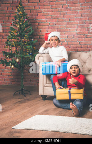 Indische Kinder feiern Weihnachten - süße kleine indische Kinder spielen, lachen und Spaß mit Geschenksets zu Weihnachten, sitzen auf dem Boden oder Sofa wi Stockfoto