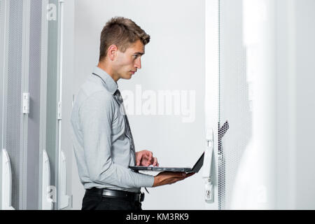 Portrait des modernen jungen Mann mit Laptop steht im Server Raum arbeiten mit supercomputer Stockfoto
