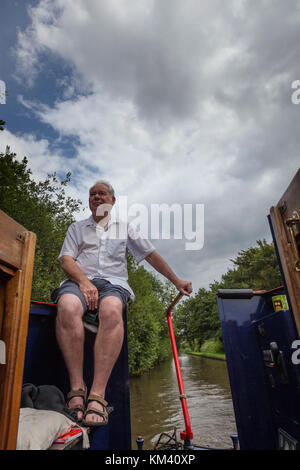 Ein Mann in kurzen Hosen und kurzen Ärmeln an der Rückseite eines 15-04, während die Lenkung mit der Lenkstange sitzt, Coventry Canal, Staffordshire, England Stockfoto