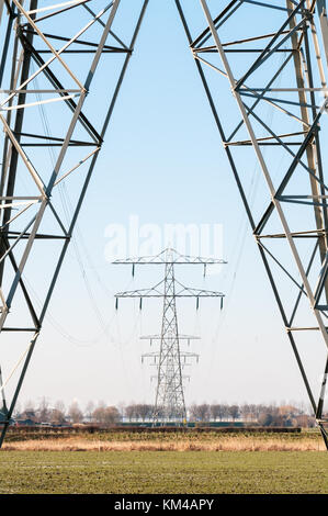 Eine Reihe von Hochspannung Strommasten stehen in einem ländlichen Gebiet und vor blauem Himmel in den Niederlanden. Stockfoto