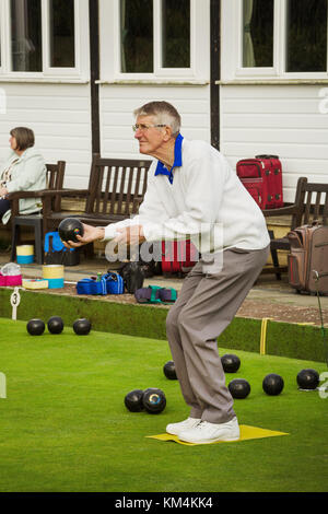 Ein grauhaariger Mann, der auf einem gelben Matte auf einem Rasen Bowling Green, biegen die Knie und Vorbereitung von seinem Schoß zu nehmen. Stockfoto