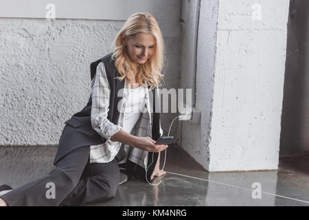 Schöne Geschäftsfrau laden Smartphone und das Sitzen auf dem Boden Stockfoto