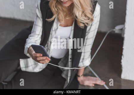 7/8 Schuß von geschäftsfrau Aufladen Handy und das Sitzen auf dem Boden Stockfoto