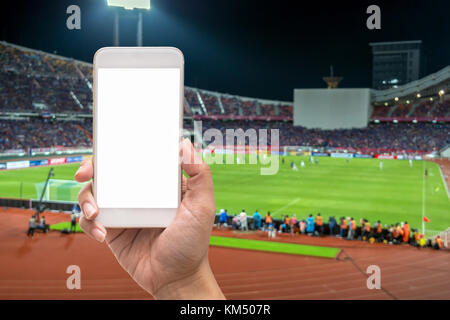 Weibliche Hand, die eine Mobile Smartphone Touchscreen auf der Fotograf, Foto bei Player in abstrakten verwischt verschwommenes Foto von Fußball-stadi Stockfoto