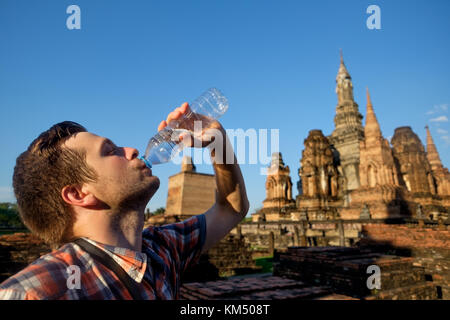 Junger Mann Durst trinkt Mineralwasser im Buddhismus Tempel in Thailand an heißen, sonnigen Wetter Stockfoto