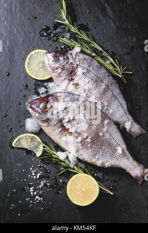Draufsicht auf Tow roher Fisch Dorade mit Rosmarin, Kalk und Eis über schwarzen Stein Hintergrund. siehe Serie Stockfoto