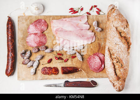 Draufsicht auf Holz Schneidebrett mit Schinken und Wurstwaren Wurst mit frischem Brot und red hot chili peppers mit Vintage Messer auf weiße Lasche serviert. Stockfoto