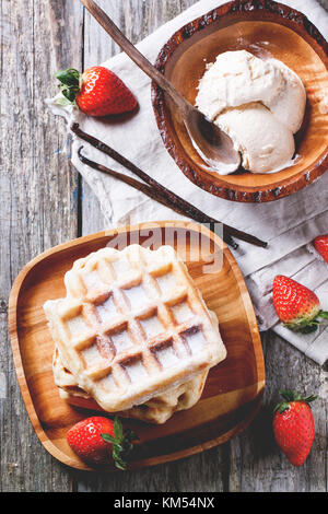 Draufsicht auf Holzplatte mit frischen Waffeln und Schüssel mit Eis, serviert mit Erdbeeren und Vanille Sticks über Holztisch. siehe Serie Stockfoto