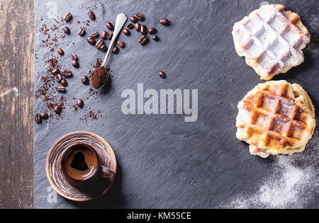 Weiße Platte mit hausgemachten belgischen Waffeln mit Zucker Pulver, Keramik Tasse Kaffee, Tl und Kaffeebohnen. Über schwarzem Schiefer als Hintergrund. mit Co Stockfoto