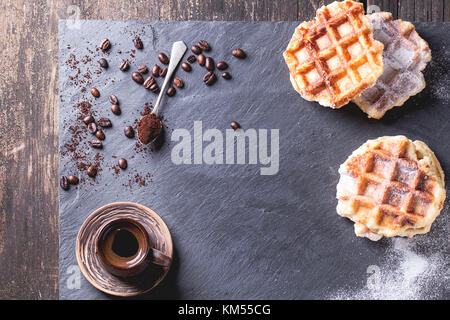 Weiße Platte mit hausgemachten belgischen Waffeln mit Zucker Pulver, Keramik Tasse Kaffee, Tl und Kaffeebohnen. Über schwarzem Schiefer als Hintergrund. mit Co Stockfoto