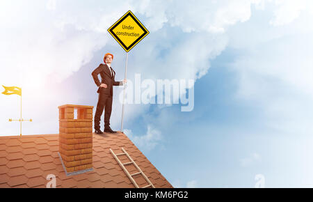 Junge Unternehmer auf Haus Ziegel Dach Holding gelbes Schild. mixed Media Stockfoto