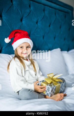 Glücklich verlassen kleines Mädchen in santa hut Holding Geschenkbox, an der Kamera schaut beim Sitzen auf dem Bett Stockfoto