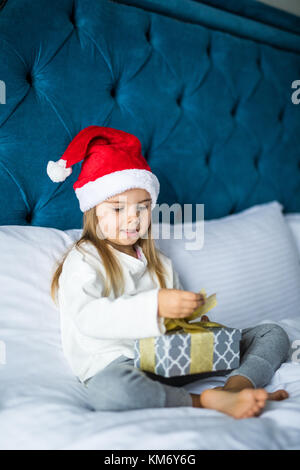 Happy kleines Mädchen in santa hut Eröffnung Geschenkbox, an der Kamera schaut mit einem Lächeln beim Sitzen auf dem Bett Stockfoto