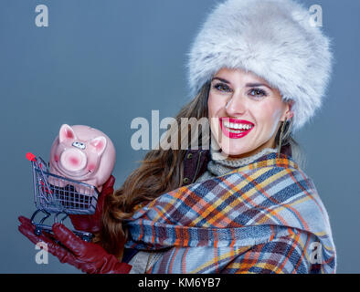 Wintersachen. Porträt der glückliche junge Frau in Pelzmütze isoliert auf kalten Blau halten Einkaufswagen mit Sparschwein Stockfoto