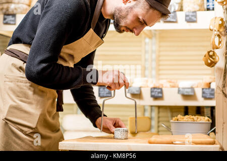 Porträt einer stattlichen Verkäufer in Uniform schneiden Käse vor dem Store Showcase voll von verschiedenen Käsesorten Stockfoto