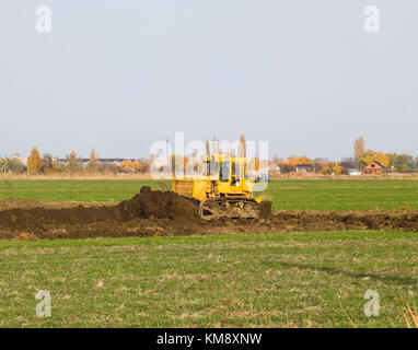 Die gelbe Traktor mit angehängtem grederom macht Boden nivellieren. Arbeiten an der Kanalisation im Feld. Stockfoto