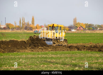 Die gelbe Traktor mit angehängtem grederom macht Boden nivellieren. Arbeiten an der Kanalisation im Feld. Stockfoto