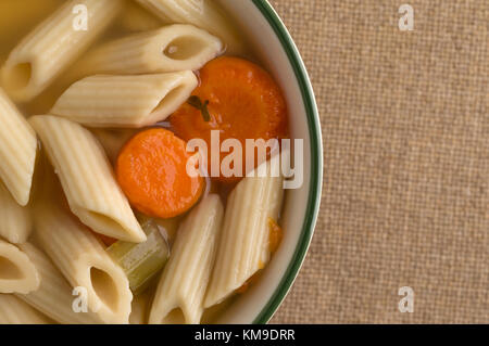 Seitenanfang schließen Blick auf eine kleine Schüssel mit Penne Pasta, Karotten und Sellerie Suppe in einer Hühnerbrühe gefüllt auf einem braunen Sackleinen Tischdecke. Stockfoto