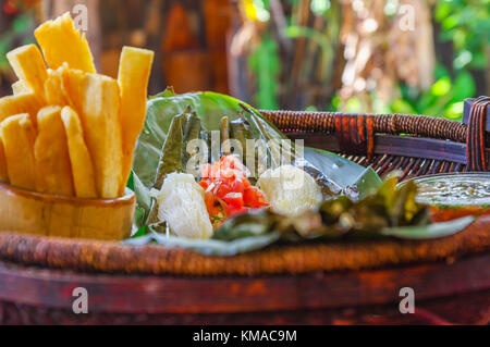 Von köstlichen typischen Amazonas Essen schließen, Fisch in einem Blatt mit Yucca und Wegerich gekocht, Schüssel Salat und gebratene Yucca, in eine hölzerne Platte über einen hölzernen Tisch serviert. Stockfoto