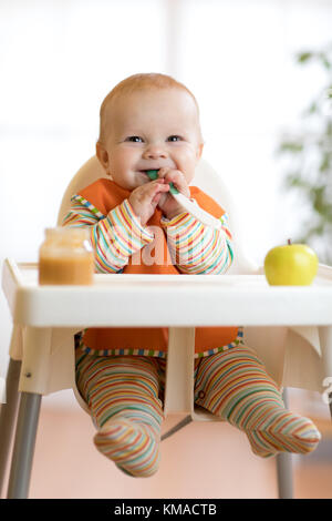 Fröhliches Baby Kind isst Essen selbst mit Löffel. Portrait von glückliches Kind Junge im Hochstuhl. Stockfoto