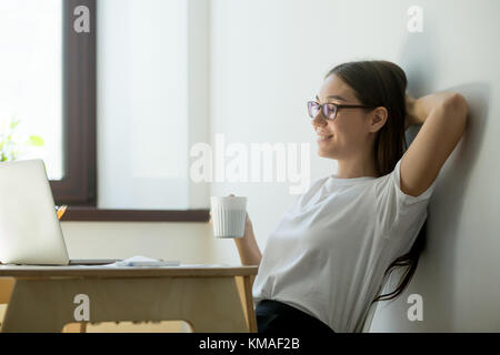 Zufriedene junge Geschäftsfrau, die Hand hinter den Kopf und trinkt Kaffee. Schöne weibliche Manager, Pause, Ruhe und lächelnd Stockfoto