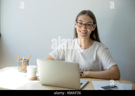 Porträt einer Millennial-Designerin, die zu Hause in einem Laptop arbeitet und über ein Notebook mit dem Internet verbunden ist. Attraktive junge Bürofrau, die lächelt und schaut Stockfoto