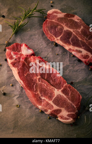 Vertikale Foto von zwei Scheiben Schweinekamm Steaks. Das Fleisch Teile mit roter Farbe und Fett auf abgenutzten Backen Blatt Papier mit Pfeffer, rosema Stockfoto