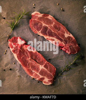 Vertikale Foto von zwei Scheiben Schweinekamm Steaks. Das Fleisch Teile mit roter Farbe und Fett auf abgenutzten Backen Blatt Papier mit Pfeffer, rosema Stockfoto