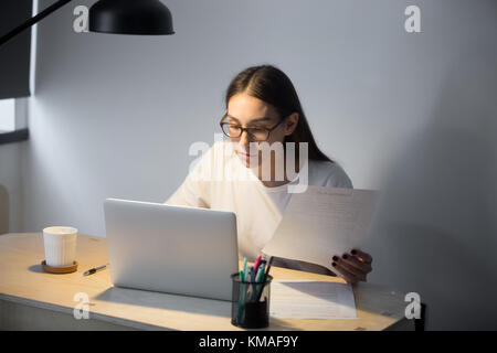 Junge casual Geschäftsfrau holding Papierdokument und Arbeiten am Laptop im Home Office in Abend. Konzentrierte weibliche Arbeitgeber prüfen Deal bei werkst Stockfoto