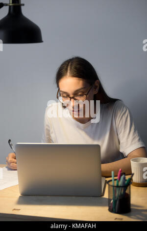 Porträt einer jungen Geschäftsfrau, die am Schreibtisch sitzt und abends am Laptop arbeitet. Müde Frau in Brille, die Überstunden schreibt und Dokumente überprüft. Vert Stockfoto