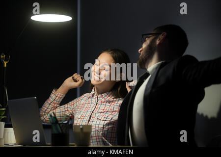 Junge positive Managern ruht im Büro nach gut getan. Lächeln, Geschäftsmann und Geschäftsfrau Entspannung und Stretching auf Stühlen in der Nacht offic Stockfoto