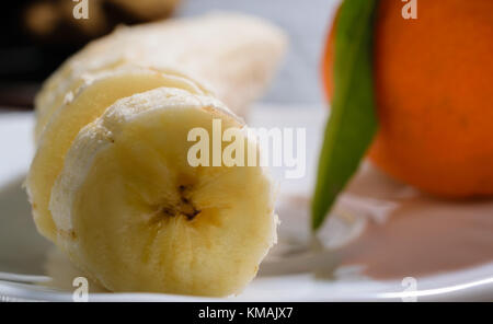 In Scheiben geschnittene Banane auf eine weiße Platte mit Mandarin Orange im Hintergrund Stockfoto