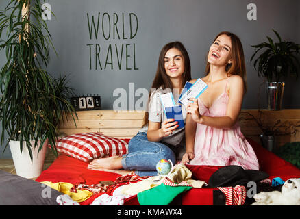Zwei fröhliche Freunde Mädchen holding Pässe und Flugtickets sitzen auf dem Bett mit Kleidung und Koffer, indoor Travel Concept Stockfoto