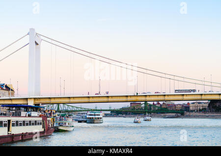 Die erste Hängebrücke der Erzsébet híd Brücke bei Sonnenuntergang über dem mit Lastkähne gefüllten Fluss Dabune in Budapest, Ungarn. Stockfoto