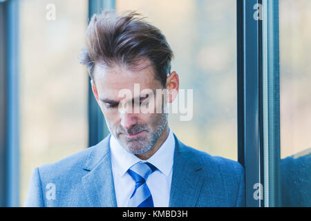 Unternehmer lehnt sich an ein Bürogebäude und schaut hinab in sein Telefon, die nicht im Bild sichtbar ist Stockfoto