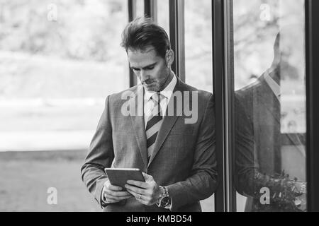 Schwarz-weiß Bild von einem Geschäftsmann lehnt sich an ein Bürogebäude mit einem Tablett in der Hand Stockfoto