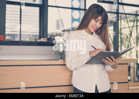 Inhaber kleiner Unternehmen holding Notebook am Zähler in Coffee Shop. asiatische Frau barista schreiben Hinweis an der Bar im Café. Food Service, Restaurant Konzept. Stockfoto