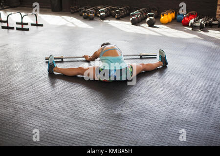 Junge Frau, die sich mit gespreizten Beinen ausdehnt, mit Langhantel im Fitnessstudio Stockfoto