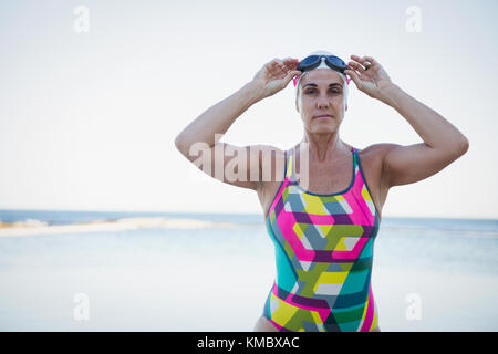 Portrait ernste, zähe reife weibliche Schwimmerin, die Schwimmbrille am Meer justiert Stockfoto