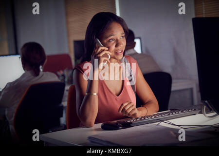 Geschäftsfrau, die spät arbeitet und im Dunkeln am Computer mit dem Smartphone spricht Büro Stockfoto