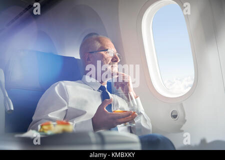 Ein älterer Geschäftsmann trinkt Whisky in erster Klasse und blickt aus dem Flugzeugfenster Stockfoto