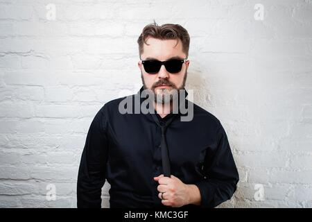 Porträt eines bärtigen ernsthaften Mannes mit Sonnenbrille, einem schwarzen Hemd und Krawatte an einer weißen Ziegelwand. Der Mann zieht sich an der Krawatte. Stockfoto