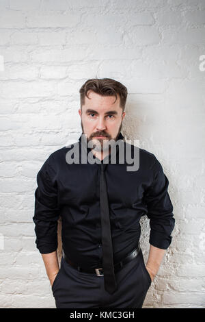 Porträt eines bärtigen ernsthaften Mannes in einem schwarzen Hemd und Krawatte an einer weißen Ziegelwand. Der Mann steht mit den Händen in den Taschen. Stockfoto