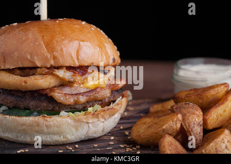 Nahaufnahme eines leckeren Burger mit Snacks in Form von Kartoffeln mit weißer Knoblauchsauce. Saftiger Burger mit Zwiebelringen, Speck und Käse auf dunklem Holz Stockfoto
